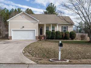 A single-story beige house on 1219 E Middleton Drive features a two-car garage, a small porch, and a neatly trimmed lawn. A black mailbox stands at the edge of the driveway leading to the sidewalk. Located in Creedmoor, trees dot the background beneath a partly cloudy sky.