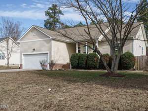 Single-story beige house with a two-car garage and a sloped roof. The front yard has a large, leafless tree and neatly trimmed bushes along the house. A wooden fence is partially visible on the right, with a clear, blue sky above.