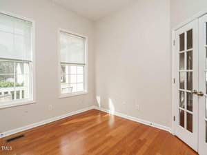 A small, empty room with light-colored walls and wooden flooring. There are two large windows with blinds, allowing natural light in. French doors with glass panels are on the right. The room appears clean and well-lit.