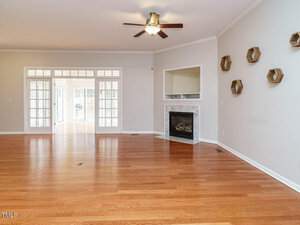 A spacious, empty living room with hardwood floors and neutral walls. There's a fireplace with a trim, a ceiling fan, and several hexagonal wall decorations. Large windows and a doorway lead to a bright adjoining room.