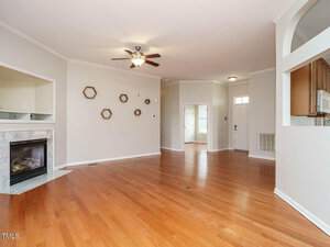 Spacious empty living room with hardwood floors, a ceiling fan, and a fireplace on the left. Hexagonal wall decor is displayed above the fireplace. An open doorway leads to the kitchen on the right, and the entrance is visible in the background.