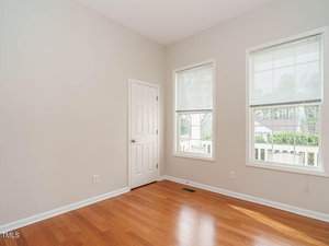 An empty room with light-colored walls and wooden flooring. It features a white door and two windows with blinds, allowing natural light to enter. Outside, part of a white fence and some greenery are visible through the windows.