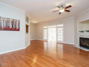 An empty living room with hardwood floors features a ceiling fan, a fireplace on the right, and a large, framed picture of a forest on the left wall. French doors lead to another room with abundant natural light. Walls are painted light beige.