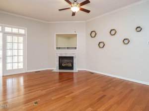 A living room with hardwood floors and light-colored walls. It features a fireplace with a white mantel and a ceiling fan with lights. Hexagonal wall accents are on the right wall. A doorway with glass panels is on the left.