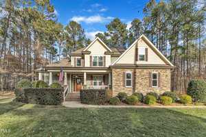 Nestled in the serene charm of Wake Forest, this two-story house at 3582 Cotton Field Court boasts a stone and beige siding exterior. A covered front porch adorned with an American flag is embraced by manicured bushes and a lush lawn, all framed by tall trees under a clear blue sky.