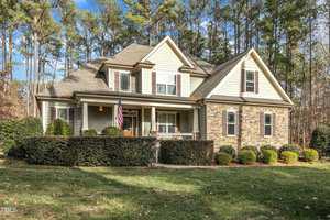 A two-story house with a mix of stone and shingle siding, surrounded by trimmed hedges and trees. An American flag hangs near the entrance, and the house features a front porch with columns. A well-maintained lawn is visible in the foreground.