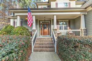 Front view of a two-story house with a welcoming porch featuring two columns and a wooden door. An American flag is displayed on the left. The porch is bordered by hedges and accessible via a short flight of brick steps.