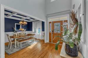 A well-lit entryway and dining room. The entryway features a wooden door and large plant. Adjacent is a dining area with a wooden table, chairs, and a chandelier. Walls are two-toned with blue and white, and a window with white curtains. Wood flooring throughout.