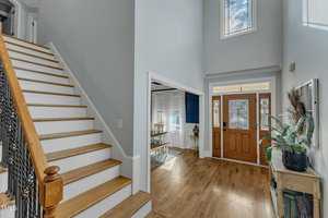 Spacious home entryway with wooden stairs, light gray walls, and a wood front door with glass panels. The hallway leads to a dining room with a blue accent wall. A potted plant sits on a small table near the entrance.