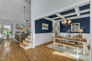The image shows a spacious open-concept living and dining area with hardwood floors. The dining room has blue walls, a coffered ceiling, and a wooden table with chairs. A staircase leads to an upper level, and a ceiling fan is visible in the living space.