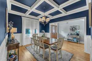 A dining room features a wooden table with six chairs, a chandelier above, and blue walls with a white coffered ceiling. A side table holds decorative items, and a window with sheer curtains lets in natural light. A door leads to a hallway.