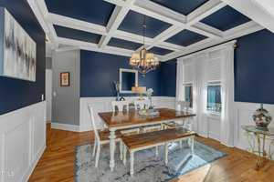 Dining room with coffered ceiling painted dark blue and white wainscoting. A wooden table with white legs is centered on a patterned rug, surrounded by white chairs and a bench. A chandelier hangs above, and a window with white curtains is on the right.