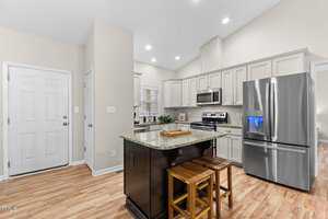 A modern kitchen with light gray cabinets and stainless steel appliances, including a refrigerator and stove. An island with a granite countertop features two wooden stools. The floor is light wood, and the walls are painted white.