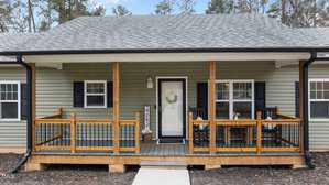 Front view of a single-story house featuring a green exterior and a covered porch. The porch has wooden railings, a white front door with a wreath, and two black chairs. The roof is gray, and there's a small pathway leading to the steps.