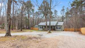 A single-story house with gray siding sits in a wooded area. There is a gravel driveway leading to the house, surrounded by trees and a wooden fence. The sky is clear and blue.