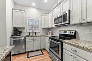A modern kitchen with light gray cabinets and granite countertops. Stainless steel appliances, including a stove, microwave, and dishwasher, are installed. A small window above the sink lets in natural light. Wood-patterned flooring is visible.