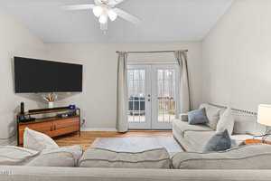 Living room with a gray sectional sofa and a wooden TV stand holding a large flat-screen TV. A ceiling fan is mounted on the white ceiling. French doors with curtains open to a view of a deck. A table lamp and some decor items are also visible.