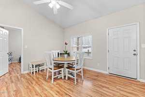 A small dining area with a round wooden table and four white chairs. There is a pink orchid on the table. The floor is light wood, and walls are beige. A ceiling fan is above, with a door and window on one wall. Another room is visible through an open door.