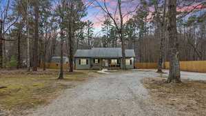 Nestled among tall trees at 4082 Old Franklinton Road, this single-story house in Franklinton features green siding and a gray roof. A gravel driveway, bordered by a wooden fence, guides you to the front entrance. The sky's pink and blue hues hint at a serene sunrise or sunset.