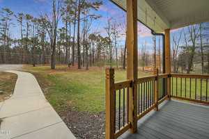 A wooden porch with black railing overlooks a yard with a paved walkway. Tall trees and a clear sky are visible in the background. The porch roof is white with decorative trim. The scene is calm and natural.