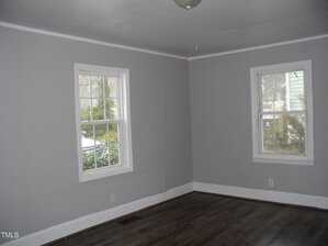 A room with two windows on adjacent gray walls, allowing natural light. The room has dark wood laminate flooring, white trim, and a central ceiling light. The windows offer a view of trees and a neighboring house.
