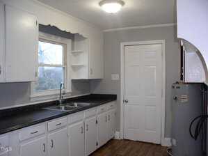 A kitchen with white cabinets and a double stainless steel sink. There is a countertop along one wall and a window above the sink. A white door and a water heater are visible. The floor is wood, and the walls are painted gray.