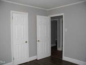 A room with gray walls and dark wood floors. Two white doors are present: one closed on the left and one partially open on the right, revealing another room. The ceiling features white molding, and a light switch is visible on the right wall.