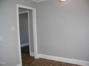 A room with light gray walls, white trim, and wood-look flooring. An open doorway leads to another room with similar decor. A light switch is visible on the left wall, and a partial view of a ceiling light is in the top right corner.