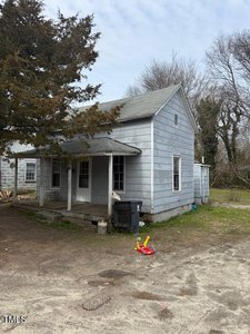 The weathered gray wooden house at 749 Southerland Street, Henderson has a porch, two small windows, and a sloped roof. A large tree towers beside it. Children's toys and a black trash bin clutter the entrance. The surrounding area is unkempt with patchy grass under an overcast sky.