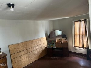 A room with white walls and a brown floor. A large, rectangular piece of plywood leans against the wall. A wooden dresser with a round mirror holds cardboard boxes. A window with sheer curtains is partially open, and a single light bulb hangs from the ceiling.