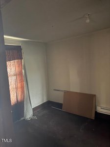 A dimly lit, empty room with a single light fixture on the ceiling. A sheer curtain covers a window on the left. A piece of wood is propped against the wall on the floor. Walls and carpet are plain and dark.