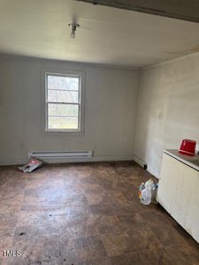 A small, empty room with a single window and a bare light bulb on the ceiling. The floor is covered with patterned vinyl tiles. A few items, including plastic jugs and a red container, rest on a white countertop to the right.