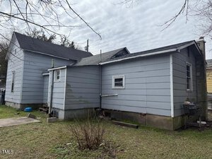 A small, single-story house with light gray siding and a dark roof is shown from a side angle. The yard is grassy with a few bushes, and a cloudy sky is overhead. A grill is visible near the structure.