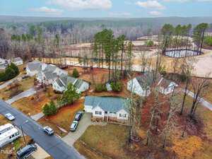 Aerial view of a residential neighborhood with several single-story houses and cars parked in driveways. The area is surrounded by trees and a cleared land area. The sky is cloudy.