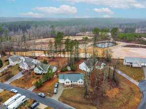 Aerial view of a suburban neighborhood with several single-story houses surrounded by trees. A small pond and a wooded area are visible in the background. The streets are curved, and a few vehicles are parked on the roads and driveways.