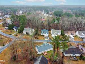 Aerial view of a suburban neighborhood with single-family homes surrounded by trees. The houses are arranged along intersecting roads. The ground appears to be wet, suggesting recent rain, and the sky is overcast.