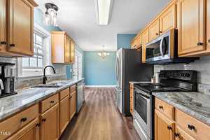 A kitchen with wooden cabinets, a granite countertop, and stainless steel appliances including a stove and microwave. A window above the sink allows natural light, and a dining area with a chandelier is visible at the far end with light blue walls.