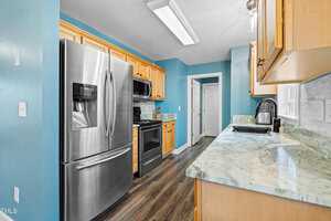 Kitchen with blue walls featuring stainless steel appliances, including a refrigerator and stove. Light wooden cabinets and green marbled countertops line the walls. A narrow space leads to a doorway with a closed white door.