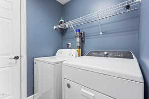 A laundry room with blue walls features a white washing machine and dryer side by side. A wire shelf above holds a detergent bottle and a wall-mounted vacuum cleaner. The room has a white door on the left.
