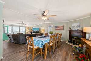 Open living and dining area with wooden flooring. A six-chair dining table covered with a blue patterned cloth is in the foreground. The room features a fireplace adorned with an American flag, large windows, ceiling fans, and a cozy sofa set.