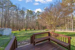 View from a wooden deck overlooking a spacious backyard. The yard features a mix of grass, trees, and shrubs, bordered by a wire fence. In the distance, a shed is visible on the left, and tall trees line the background under a partly cloudy sky.