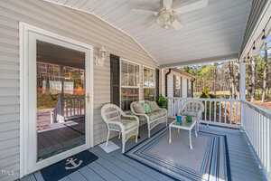 A covered porch features wicker furniture with cushions, a small table, and a ceiling fan. The area is enclosed by a white railing and overlooks a yard with trees. The porch has gray flooring and a welcome mat near the glass door.