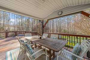 A wooden deck with a rectangular dining table and eight chairs is under a covered area. Cushions are on the chairs, and string lights hang overhead. The deck overlooks a grassy yard with trees in the background.