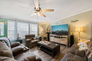 A cozy living room with two couches and an armchair facing a flat-screen TV. The room features a ceiling fan, large windows with natural light, a wooden coffee table, and wall decor including a fish collection and a "Home" sign.