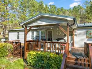 A wooden deck with a covered porch attached to a gray house. The deck has railings and stairs leading down to a green lawn. There are string lights hanging from the porch roof. Trees are visible in the background under a clear blue sky.