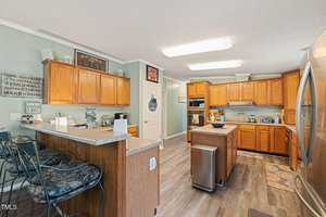 A kitchen with wooden cabinets, stainless steel appliances, and a central island. Two cushioned bar stools are at the counter. Wall decor includes signs with text. The floor is hardwood, and overhead lighting illuminates the space.