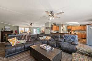 A spacious living room with a large sectional sofa facing a wooden coffee table. The room features ceiling fans, wood flooring, and opens into a kitchen with wooden cabinets. Decor includes art pieces, pillows, and a decorative clock.