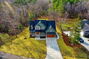 Aerial view of a large, two-story house with a sloped roof and a three-car garage. The house is surrounded by a grassy yard and bordered by a dense wooded area. There is a wide driveway leading up to the front of the house.