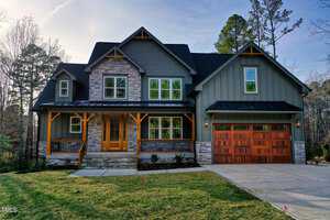 Located at 414 S Sb Harrison Lane in Henderson, this charming two-story house boasts gray siding and stone accents. It features a welcoming wooden porch and a double garage with wooden doors. The lush yard is dotted with trees against the backdrop of a clear sky.