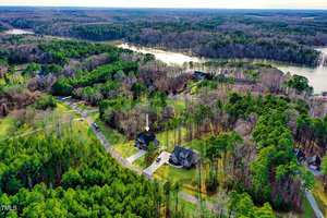 Aerial view of a residential area surrounded by green trees. Two houses are visible with driveways, and a river winds through the landscape in the background. An arrow points to one of the houses. The sky is partly cloudy.